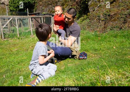 Padre asiatico padre padre con due figli ragazzi fratello Guardare seduti fuori in primavera tenente toddler in Carmarthenshire Galles REGNO UNITO KATHY DEWITT Foto Stock