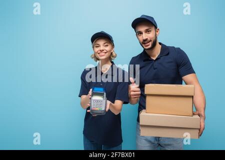 felice uomo di consegna arabo che mostra il pollice in su vicino collega con terminale di pagamento su blu Foto Stock