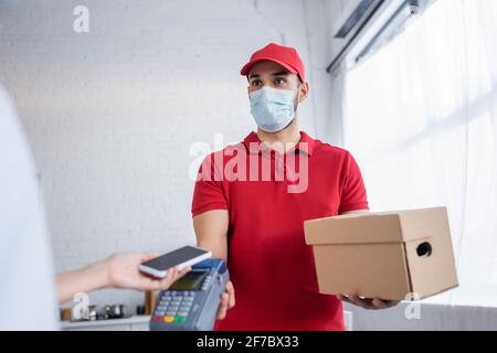 corriere musulmano in maschera medica che tiene terminale di pagamento vicino a donna con lo smartphone in primo piano sfocato Foto Stock