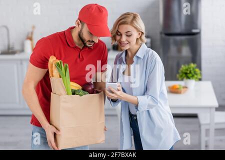 donna sorridente che mostra smartphone al corriere arabo che tiene il sacchetto di carta con cibo fresco Foto Stock