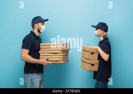 vista laterale dei corrieri interrazziali in maschere mediche che tengono pacchetti su blu Foto Stock