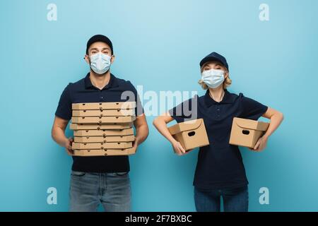 corrieri interrazziali in maschere protettive contenenti scatole per pizza e cartone confezioni su blu Foto Stock