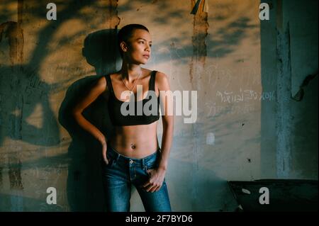 Giovane donna asiatica con capelli corti e reggiseno sportivo e jeans blu in piedi contro una parete con graffiti e ombre, guardando a lato nella luce. Foto Stock