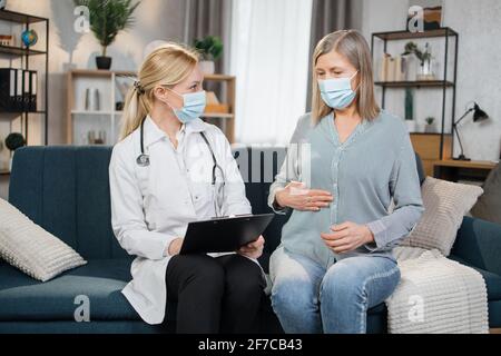 La donna anziana che indossa una maschera protettiva parla con un medico a casa e si lamenta del mal di stomaco. Donna anziana preoccupata che parla con il suo medico generale che la visita a casa durante l'epidemia di virus Foto Stock