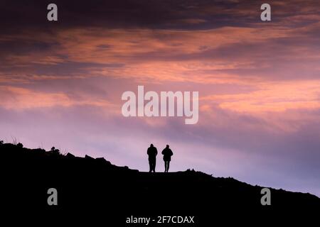 Due persone si stagliavano contro un cielo colorato che camminava attraverso il promontorio sulla costa di Newqay in Cornovaglia. Foto Stock