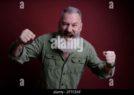 Uomo maturo con gesto difensivo pugni, uomo arrabbiato e sconvolto su sfondo rosso, pronto a combattere, paura di problemi Foto Stock