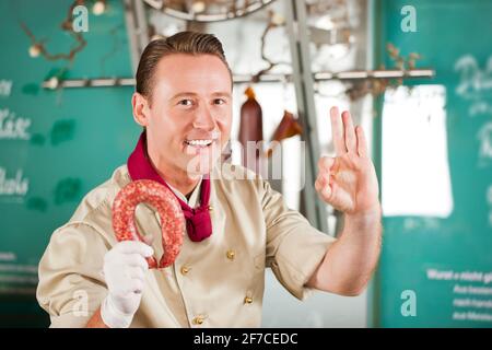 Ritratto del macellaio che mostra un segno positivo mentre si tiene la salsiccia affumicata Foto Stock