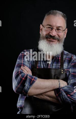 Ritratto di bel grembiule in pelle vestito uomo maturo isolato su sfondo nero, operaio caucasico con barba sorridente Foto Stock