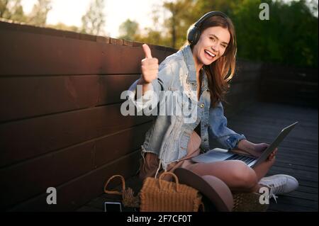 Giovane ragazza sorridente felice che lavora con il computer portatile per strada ascolta la musica sulle cuffie e mostra il pollice in su. Concetto di apprendimento a distanza Foto Stock