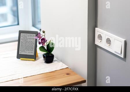 Gruppo di prese elettriche europee bianche e un interruttore situato su una parete grigia in una cucina moderna e luminosa con fiore e e-libro sul tavolo Foto Stock