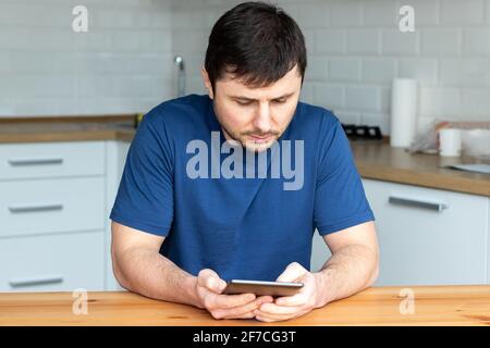 Un bell'uomo bearded in una T-shirt blu si siede dietro a. tavola di legno e legge un libro elettronico contro un offuscato sfondo di una cucina moderna Foto Stock