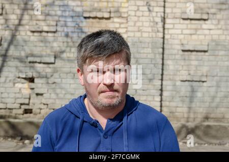 Ritratto di un uomo maturo con capelli grigi corti contro una parete di mattoni. Un uomo di mezza età in una camicia blu con cappuccio guarda nella fotocamera. Esterno. Giorno. Foto Stock