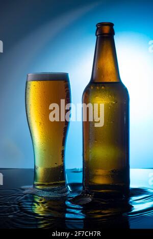 bottiglia di birra versata in acqua su una bottiglia blu di birra con un bicchiere di birra schiumosa su sfondo blu, sull'acqua Foto Stock