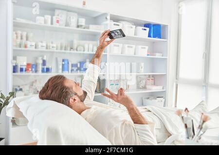 Giovane uomo sorridente e facendo selfie mentre attende per cosmetic procedura in clinica di cosmetologia estetica Foto Stock
