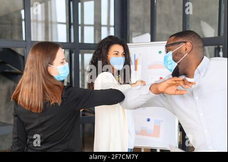 Diverse due donne e l'uomo, colleghi in maschere facciali di protezione saluto urtare gomiti sul posto di lavoro, donna collaboratori in coperture facciali proteggere da COVID-19 coronavirus in ufficio, concetto di assistenza sanitaria Foto Stock