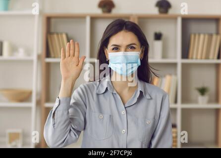 Giovane donna caucasica dipendente remoto in maschera agitando mano a. ritratto della fotocamera Foto Stock