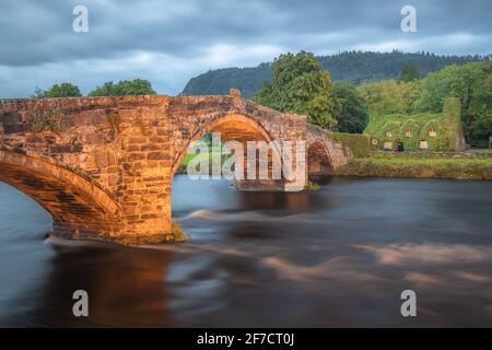 Vecchio ponte di ispirazione romana Pont Fawr attraverso il fiume Conwy verso una casa di pietra coperta di edera del 15 ° secolo nel villaggio gallese di Llawnrst, Galles del Nord Foto Stock