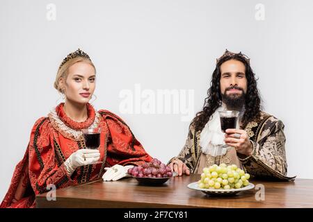 coppia storica interrazziale in corone con calici di vino rosso mentre si siede a tavola con uve isolate su bianco Foto Stock