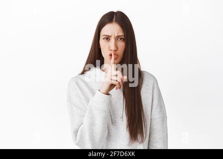 Ragazza seria dire a Fai silenzio. Donna che grida e fa il segno del tabù, tenga segreto, si comporti, silenzi prego, levandosi in piedi contro sfondo bianco Foto Stock