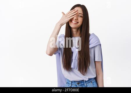 Bella giovane donna bruna sorridente, che copre un occhio e guardare adorabile alla macchina fotografica, in piedi in t-shirt su sfondo bianco Foto Stock