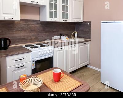 Interno cucina beige con set cucina bianco e elettrodomestici, tavolo con utensili. Foto Stock