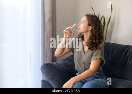 Giovane donna riccia sente sete a casa, bevendo acqua pura fresca da vetro trasparente, ripristina l'equilibrio dell'acqua, si prende cura della salute. Concetto di idratazione Foto Stock