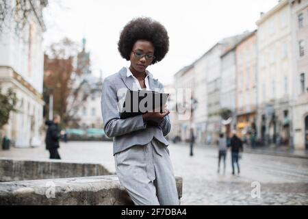 Donna africana focalizzata in elegante abito da lavoro seduto con appunti sulla strada della città. Donna di affari in occhiali che scrivono le note di lavoro all'aperto. Foto Stock