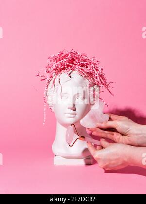 Rulli di pietra di giada del viso e massaggio di gua sha per la piantatrice trendy di testa di gesso Venus con cervello di carta su sfondo rosa. Copia in gesso dell'antica statua Foto Stock