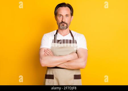 Foto di un uomo serio professionista che tiene le mani piegate grembiule isolato su sfondo giallo Foto Stock