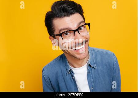 Primo piano di un simpatico e allegro ragazzo caucasico con occhiali e camicia in denim, in piedi su sfondo arancione isolato, guardando direttamente nella fotocamera, sorrisi toothy Foto Stock