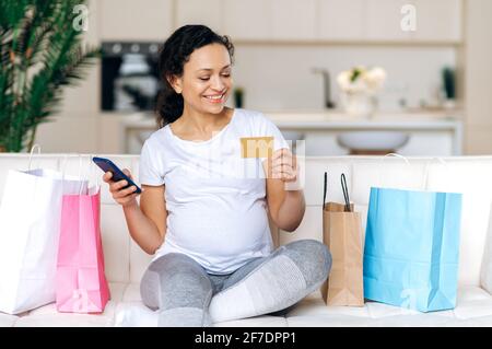Concetto di shopping online. Attraente donna di corsa mista felice incinta, seduta sul divano, shopping online, tenendo la carta di credito e il telefono cellulare, facendo il pagamento online, sorridendo Foto Stock