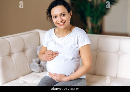 Bella donna di razza mista, adulta felice incinta, indossando una t-shirt bianca di base, seduta a casa sul divano, stracciandola delicatamente con le mani, guardando la macchina fotografica, sorridendo felice Foto Stock