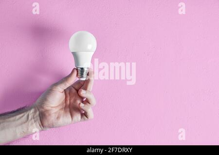 Mano di un giovane adulto che tiene una lampadina a risparmio energetico contro lo sfondo rosa della parete. Concetto di creatività, efficienza e sostenibilità. Foto Stock