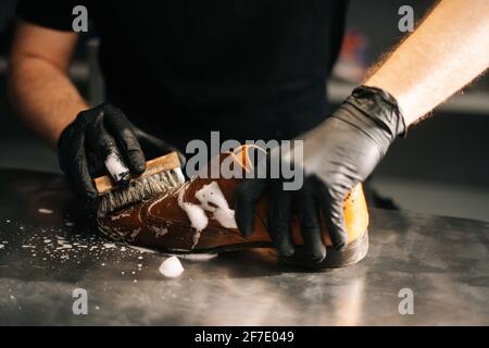 Mani ravvicinato di calzolaio irriconoscibile in guanti neri applicando schiuma detergente su vecchie scarpe in pelle marrone chiaro. Foto Stock