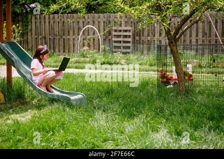 Una bambina si siede sul playset in giardino con il computer nella scuola virtuale Foto Stock