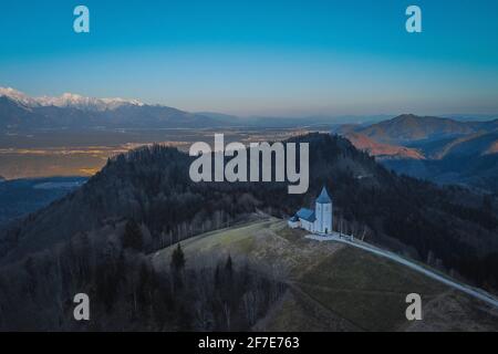 Foto di un drone aereo della graziosa e fantastica chiesa di Saint Primoz su una piccola collina con montagne sullo sfondo al tramonto, Jamnik villaggio, Slovenia, UE Foto Stock