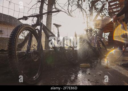 Persona che pulisce mountain bike con l'aiuto di un getto d'acqua. Foto panoramica sognante della pulizia delle biciclette, retroilluminazione forte e soleggiata. Foto Stock