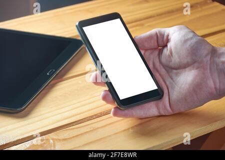 Mano di un giovane adulto che tiene il telefono cellulare nero con schermo vuoto bianco su un tavolo rustico in legno. Tecnologia, comunicazione, lavoro da casa. Foto Stock