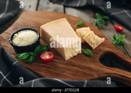 Concetto di cucina Italiana. Tagliere, pezzi di formaggio parmigiano, formaggio grattugiato in ciotola nera, tomi di ciliegia, rami di foglie di basilico, torce da cucina Foto Stock