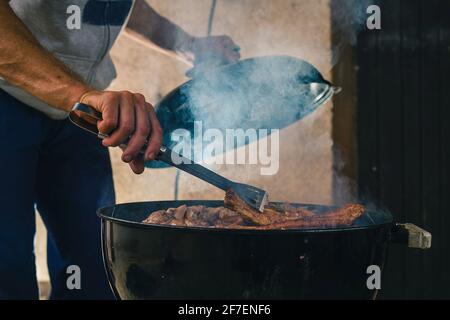 Uomo che apre un barbecue fumatore o un forno a carbone, per girare kebab o costolette di ricambio grigliare a caldo. Fumo visibile che sale dalla griglia Foto Stock
