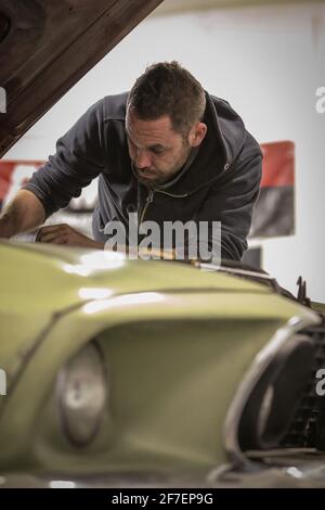 Un giovane sta raggiungendo sotto il cofano di una vecchia auto d'epoca che fissa il motore. Uomo che lotta per riparare il motore di una macchina del muscolo verde. Foto Stock