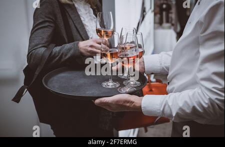 Persone che prendono bicchieri di vino rosato da un vassoio tenuto da un cameriere o da un ristoratore professionista. Catering con vino in un evento aziendale all'interno. Foto Stock