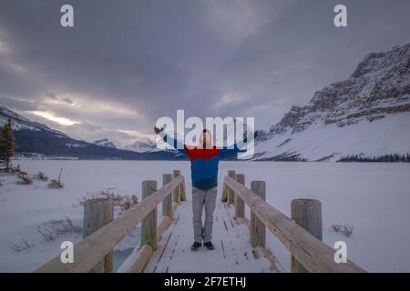 L'uomo sta alzando le mani sul ponte di legno del lago Bow ad Alberta, Canada. Vista frontale di un uomo in un punto epico sul lago Bow di prima mattina. Foto Stock