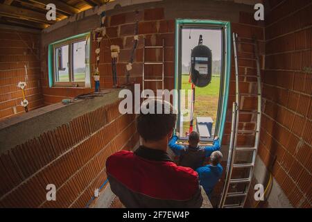 Tre lavoratori maschi che installano una moderna finestra di vetro in una nuova casa che viene costruita utilizzando un sollevatore idraulico o una gru montata su un camion. Installazione moderna Foto Stock