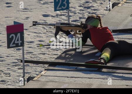 La corsa femminile del biathlon sta sdraiandosi sul terreno e sta puntando il suo fucile. Donna biatleta su un raggio di tiro, sparando sdraiato. Foto Stock