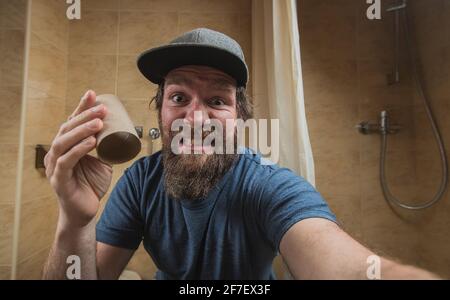 Uomo hipster con una faccia distretta o disturbata come tiene un rotolo vuoto di carta igienica in bagno. Uomo spaventato di esaurire la carta igienica. Foto Stock