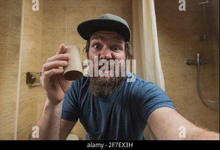 Uomo hipster con una faccia distretta o disturbata come tiene un rotolo vuoto di carta igienica in bagno. Uomo spaventato di esaurire la carta igienica. Foto Stock