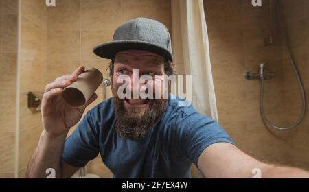 Uomo hipster con una faccia distretta o disturbata come tiene un rotolo vuoto di carta igienica in bagno. Uomo spaventato di esaurire la carta igienica. Foto Stock