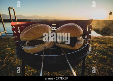 Grigliare gli hamburger su un piccolo grill portatile in metallo al carbone attivo Le rive del Lac Leman in Svizzera durante il primo inverno serata con tramonto Foto Stock