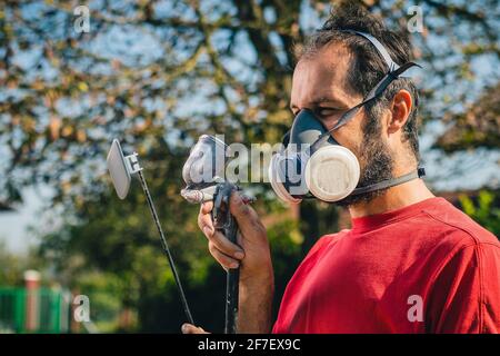 Pittore dilettante in maglia rossa e maschera facciale con una pistola a spruzzo per dipingere un oggetto di plastica o una parte in un giardino domestico. Profilo di un pittore con un mq Foto Stock